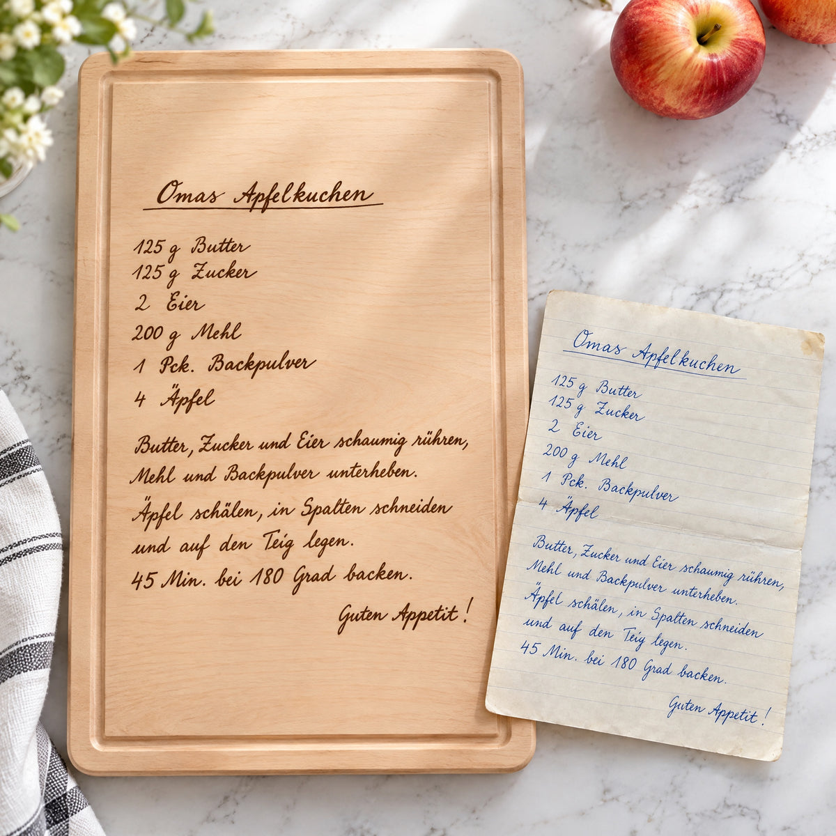 Graviertes Rezept-Schneidebrett mit Omas Apfelkuchen auf hellem Marmor neben dem handgeschriebenen Originalrezept